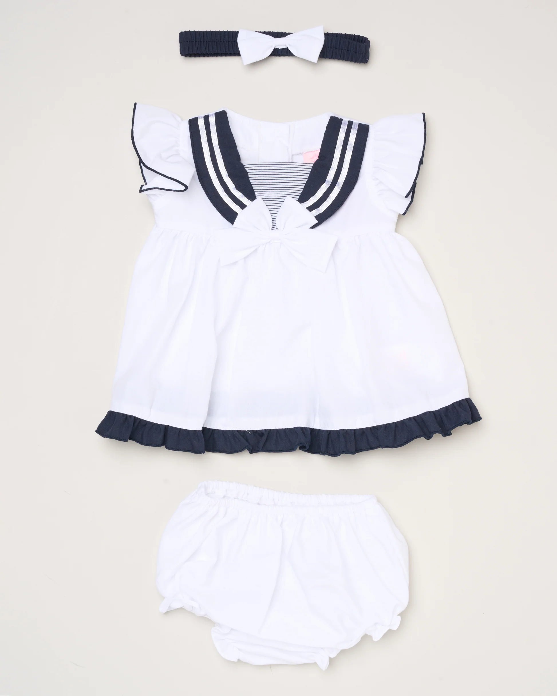 The Baby Girls Dress, Pant & Headband Set includes a white sailor-style dress with navy trim and ruffles, matching bloomers, and a navy headband with a white bow—all beautifully displayed on a light background.