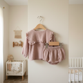 The Girl's Soft Infant Clothes Bread Ruffle-collar Lapel Top Pants Two Piece Set in mauve—featuring a ruffled long-sleeve top and matching cotton pants—hangs on a nursery wall, with a crib, teddy bear, and toys in the background.