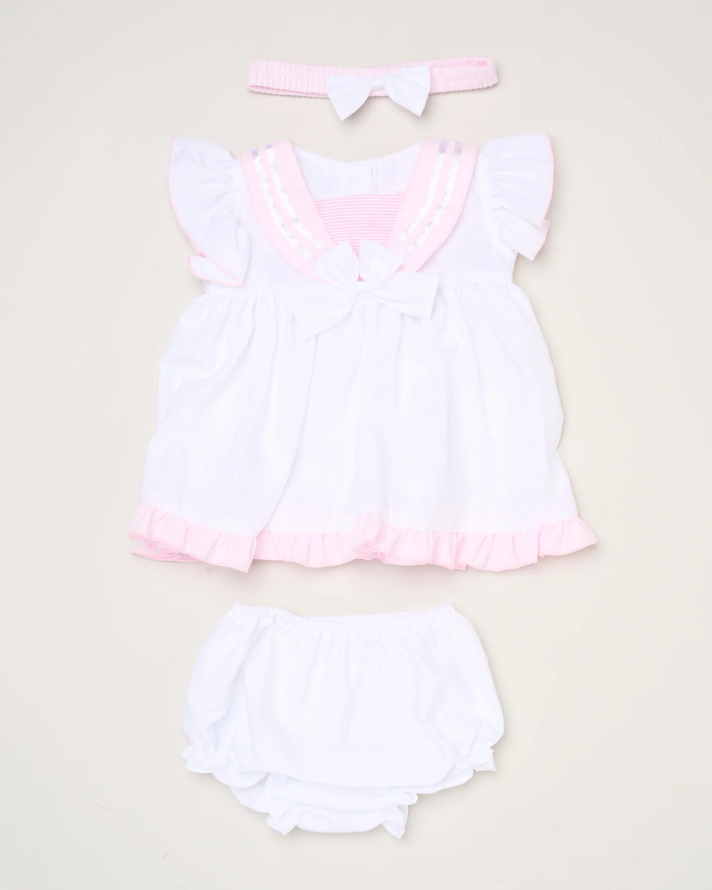 The Baby Girls Dress, Pant & Headband Set features a white dress with pink trim and ruffles, a matching white diaper cover, and a pink headband with a white bow, beautifully displayed on a light background.